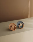 The FRIGG Daisy Silicone Pacifier 2-Pack by FRIGG features two medical grade silicone pacifiers—one soft peach, one muted gray—resting on a beige surface in warm, natural light for ultimate baby comfort.