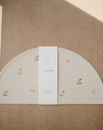 A beige, semicircular mushie Silicone Placemat made from food-grade silicone and decorated with cherry illustrations sits on a brown textured surface, wrapped in a white mushie band and lit by a diagonal ray of light.