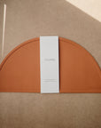 A rust-colored, half-circle Silicone Placemat by mushie with a white paper label is displayed on a tan surface under soft natural light.
