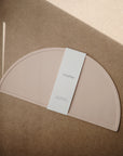 A light brown, semicircular Silicone Placemat by mushie made from food-grade silicone, with a white label reading "mushie" wrapped around the center, sits on a tan surface with sunlight casting shadows.