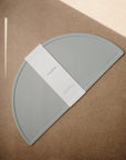 A light gray, semi-circular mushie Silicone Placemat made from food-grade silicone rests on a brown surface. Sunlight casts diagonal shadows as the placemat sits partially wrapped in white paper packaging.
