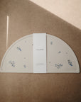 The mushie Silicone Placemat, in beige food-grade silicone with small blue floral prints and a semicircular shape, lies flat on a brown surface. Sunlight casts a diagonal shadow across its center, highlighting the white mushie label.