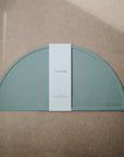A half-circle, sage green Silicone Placemat by mushie, made of food-grade silicone with the mushie logo embossed in the corner, is partially wrapped in a white band labeled mushie and displayed on a tan surface.