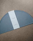 A blue, semi-circular mushie Silicone Placemat made from food-grade silicone is shown on a brown surface, wrapped with a white paper label at the center.