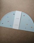 A light blue, semi-circular Silicone Placemat by mushie, made from food-grade silicone with colorful confetti shapes, sits on a brown surface. A white paper band with the mushie logo wraps around the center of the mat.
