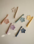Seven mushie Flower Training Toothbrushes in pastel shades—pink, beige, blue, green, yellow, purple, and navy—with matching flower-shaped holders are scattered on a light beige surface. The toothbrushes are made of silicone.