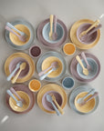 A top-down view of nine sets of mushie Dinnerware Cups and matching pastel plates, bowls, forks, and spoons in soft yellow, blue, and mauve tones. Non-toxic plastic dinnerware is arranged in a grid on a light surface.