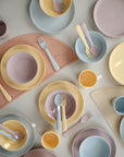 A selection of mushie Round Dinnerware Bowls in pastel pink, yellow, lavender, and blue are neatly arranged on a light surface with a semi-circular placemat. These BPA-free plastic bowls are perfect for children.