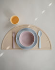 A minimalist kids’ tablescape features mushie Dinnerware Cups (non-toxic), a beige semicircle placemat, a pink fork, a blue spoon, and a pink bowl stacked in a blue plate—all arranged neatly on a light surface.
