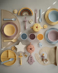 A neatly arranged mushie silicone baby feeding set features a toddler-friendly plate, bowl, utensils, the mushie Snack Cup, pacifiers, teethers, a rainbow, and fun pastel shapes on a light background.