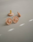Two beige FRIGG Daisy Silicone Pacifiers, featuring daisy-shaped shields with scalloped edges, rest on a light gray surface as soft natural light creates dotted shadows across the scene.