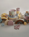 A set of pastel-colored children's tableware and toys—including bowls, cups, stacking rainbows, a bunny teether, pacifier, mushie’s Flower Training Toothbrush, rockets, a star, and a pretzel—arranged on a neutral surface.