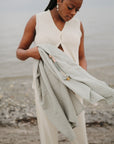 A woman stands on the beach holding the mushie Organic Cotton Muslin Swaddle Blanket with beaded pacifier clips. She wears a cream sleeveless top and pants, with the shoreline and water in the background.