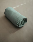 The mushie Extra Soft Bassinet Muslin Crib Sheet, in light green, is neatly rolled on a beige surface with soft lighting that highlights its cozy, breathable texture—ideal for gentle baby bedding.