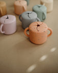 Several mushie Snack Cups with handles and slotted lids are arranged on a beige surface; the focus is on a speckled, peach-colored silicone, spill-proof toddler Snack Cup in the foreground.