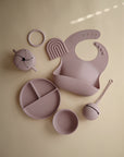 The mushie Snack Cup in mauve is a silicone feeding essential for toddlers, featuring a spill-proof design and matching bib, divided plate, bowl, sippy cup, spoon, teether, and rainbow teething toy—perfectly arranged on a beige background.