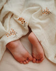 Close-up of a baby’s feet peeking out from under a mushie Organic Cotton Muslin Swaddle Blanket, featuring small orange and brown floral patterns on soft, white fabric.