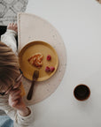 A child eats a heart-shaped toast from a yellow plate with raspberries at a white table, using a fork and cup. The table features the mushie Silicone Placemat, made of food-grade silicone.