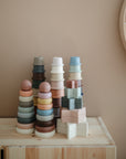 Pastel-colored baby cups and blocks are displayed on a light wooden surface with the mushie Stacking Rings Toy. A round shelf holding small items appears to the right, all set against a beige wall.