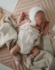A newborn in a white bonnet, romper, and socks lies on a beige quilt with a mushie Organic Cotton Muslin Swaddle Blanket and neutral-toned blankets nearby, holding a pacifier and surrounded by a soft toy.