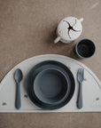 A mushie Dinnerware Cups set is displayed with a gray children’s plate, bowl, spoon, fork on a semi-circular placemat with rainbow patterns. A non-toxic kids cup and white snack cup with a star opening sit above on a brown surface.
