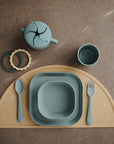 A mushie Dinnerware Cups set for kids features non-toxic cups, blue bowls, a snack container, yellow placemat, fork, spoon, and three teething rings neatly arranged on a brown surface.