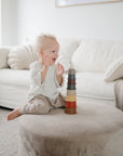 A smiling toddler sits on a round ottoman in a cozy living room, happily clapping hands while eyeing a stack of mushie Stacking Cups Toy. Soft furnishings and a white couch create a warm, inviting background.
