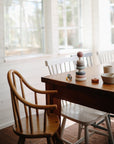 A wooden dining table with white chairs and a high chair is bathed in sunlight. On the table are stacked cups, bowls, and the mushie Stacking Rings Toy. Large windows brighten the cozy minimalist decor with natural light.