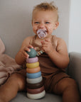 A smiling baby in a brown outfit sits in a chair, holding the mushie Stacking Rings Toy in one hand and a pacifier in the other, with a light pink blanket draped beside them.