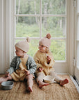 Two young children in knit hats and sweaters sit barefoot on a rug by large windows, holding mushie Square Dinnerware Bowls. They look out at the leafy yard as soft natural light fills the room.
