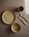 A set of mushie Dinnerware Cups and matching beige children’s dishes—including a plate, two bowls, a cup, fork, and spoon—is neatly arranged on a brown surface. The plate and one bowl have "mushie" embossed on them.