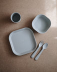 A set of light gray mushie Dinnerware Cups and dishes for kids—including plate, bowl, cup, fork, and spoon—is neatly arranged on a brown surface. The non-toxic pieces have "mushie" embossed on the plate and bowl.