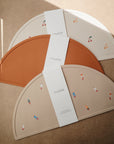Three semi-circular mushie Silicone Placemats in beige, tan, and rust (one solid rust, others with fruit and fish designs), each wrapped in a white band. Made from food-grade silicone for safe, easy mealtime cleanup.