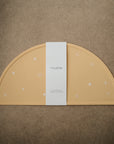 A mustard yellow, semi-circular mushie Silicone Placemat made from food-grade silicone features white starburst designs and rests on a brown surface, with a white paper band displaying the mushie brand name wrapped around its center.