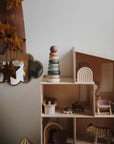 A mushie Stacking Rings Toy sits beside a wooden dollhouse decorated with non-toxic toy furniture, a small toy tiger, pastel arches, a cloud-shaped mirror, and hanging star decorations.