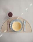 A minimalist table setting showcases mushie's Round Dinnerware Bowl, paired with a beige half-circle placemat, yellow baby bowl, light blue spoon, pink fork, and mauve cup—all BPA-free and beautifully arranged under soft light.