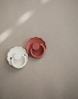 The FRIGG Daisy Silicone Pacifier 2-Pack by FRIGG includes one cream and one terracotta pacifier with daisy-shaped shields, each featuring a medical grade silicone nipple for safety and comfort.
