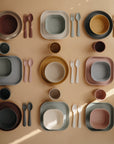 A flat lay of mushie Dinnerware Cups among matching plates, bowls, forks, and spoons in pink, mustard, green, gray, and brown hues arranged on a beige surface in a stylish grid pattern. All pieces are non-toxic and perfect for kids.