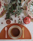 A festive table setting with decorative plates, glasses, evergreen branches, acorns, a pomegranate, and a child's place set featuring the mushie Silicone Placemat and matching food-grade silicone bowl, cup, spoon, and fork in warm earthy tones.
