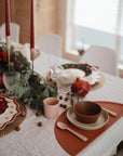 A festive table is set for the holidays with plates, glassware, red candles, greenery, and reindeer. In the front, a brown and pink kids’ place setting features mushie’s non-toxic Dinnerware Cups and plastic cups.