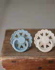 The FRIGG Andersen Fairytale Silicone Pacifier 2-Pack by FRIGG, featuring light blue and cream pacifiers with scalloped edges and heart-shaped cutouts, rests on a rustic wooden surface.