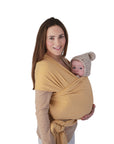 A woman smiles while carrying a baby in a mushie Baby Wrap made from organic cotton. The baby, wearing a pom-pom knitted hat, rests snugly against her chest against a plain white background.