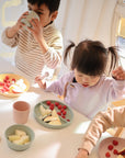 Three children sit at a table, enjoying apple slices and raspberries from mushie Silicone Suction Plates. One sips from a cup as soft, natural light fills the scene—making baby feeding both simple and joyful with mushie’s thoughtful design.