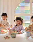 Three young children smile as they eat fruit with the mushie Dinnerware Fork and Spoon Set. Pastel cups and plates decorate the bright room, while a colorful stained-glass window shines behind their white table.