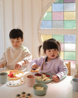 Three young children sit at a table with plates of fruit and pastel cups, using mushie Silicone Feeding Spoons in a sunlit room with stained glass windows in the background.