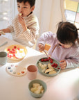 Two young children sit at a white table eating fruit from colorful plates. One uses a mushie Silicone Feeding Spoon with sliced pear and raspberries, while the other eats raspberries by hand. Cups and extra fruit are also on the table.