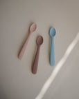 Mushie Silicone Feeding Spoons—three food-grade spoons in pastel pink, mauve, and light blue—are arranged in a triangle on a light surface, illuminated by a diagonal streak of light. Perfect gentle baby spoons.