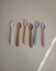 Six mushie Silicone Feeding Spoons in pastel food-grade silicone—white, brown, light pink, peach, light blue, and lavender—are arranged side by side on a light beige background.