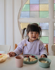 A young child smiles in her light purple sweater at a table set with mushie Silicone Feeding Spoons, pastel bowls, and cups of fruit, while colorful stained glass glows behind her.