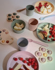 Mushie Dinnerware Cups in soft pink, green, and beige tones are displayed with colorful silicone kids’ plates, bowls, spoons, and shape sorters, paired with raspberries and apple slices on a white surface.